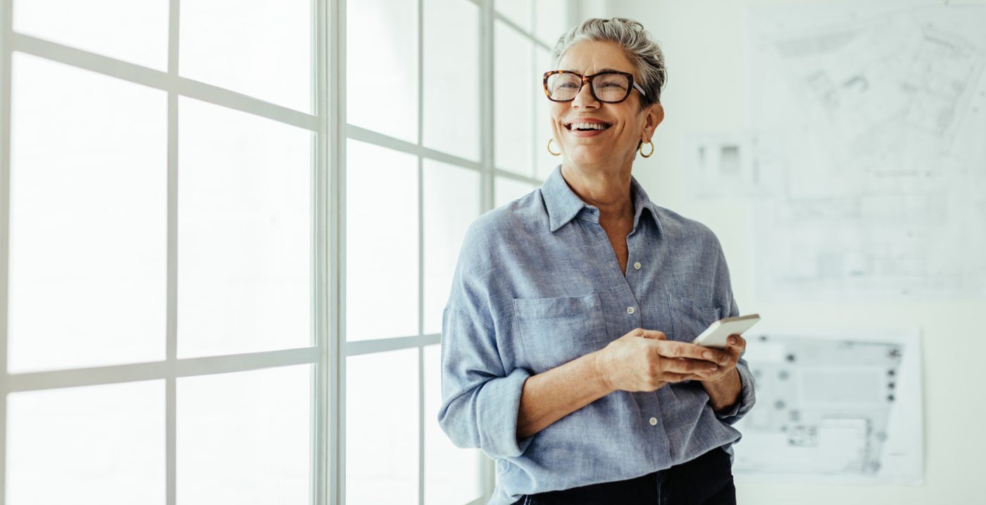 A smiling woman with short grey hair and glasses holding a phone beside a bright window