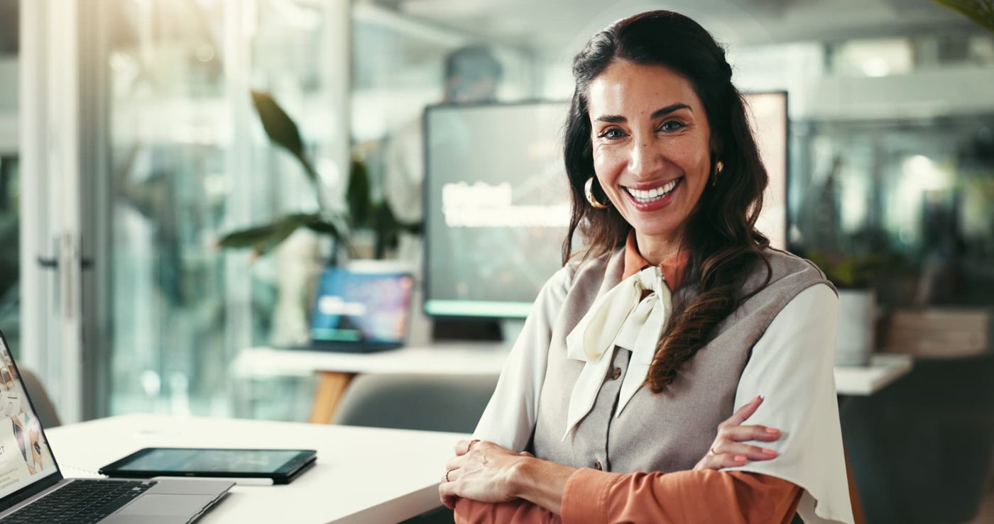 A smiling professional woman in a waistcoat and orange blouse standing in a bright office